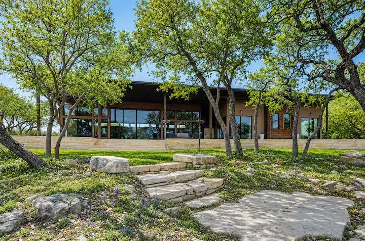 La Paz exterior — Hill Country home framed by mature live oaks, limestone walls, and a winding stone path through native wildflowers